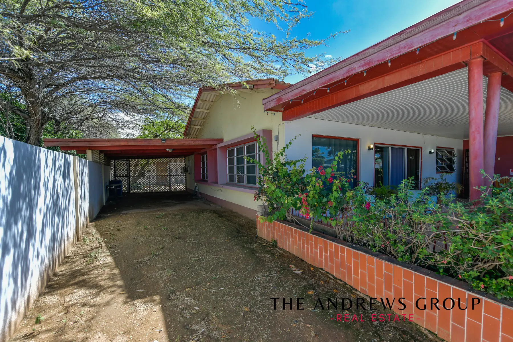 The carport side today: a covered carport with a lattice gate, a bare earth drive, red veranda posts and fascia, bougainvillea over a red brick planter.