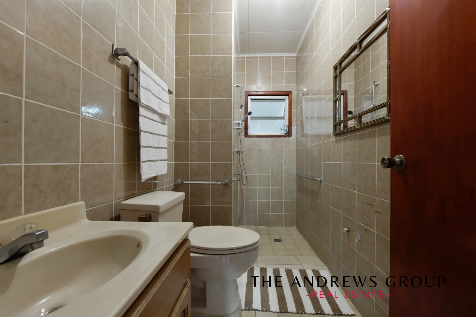 A bathroom tiled floor to ceiling in beige, with a walk-in shower, toilet, vanity and mirror.