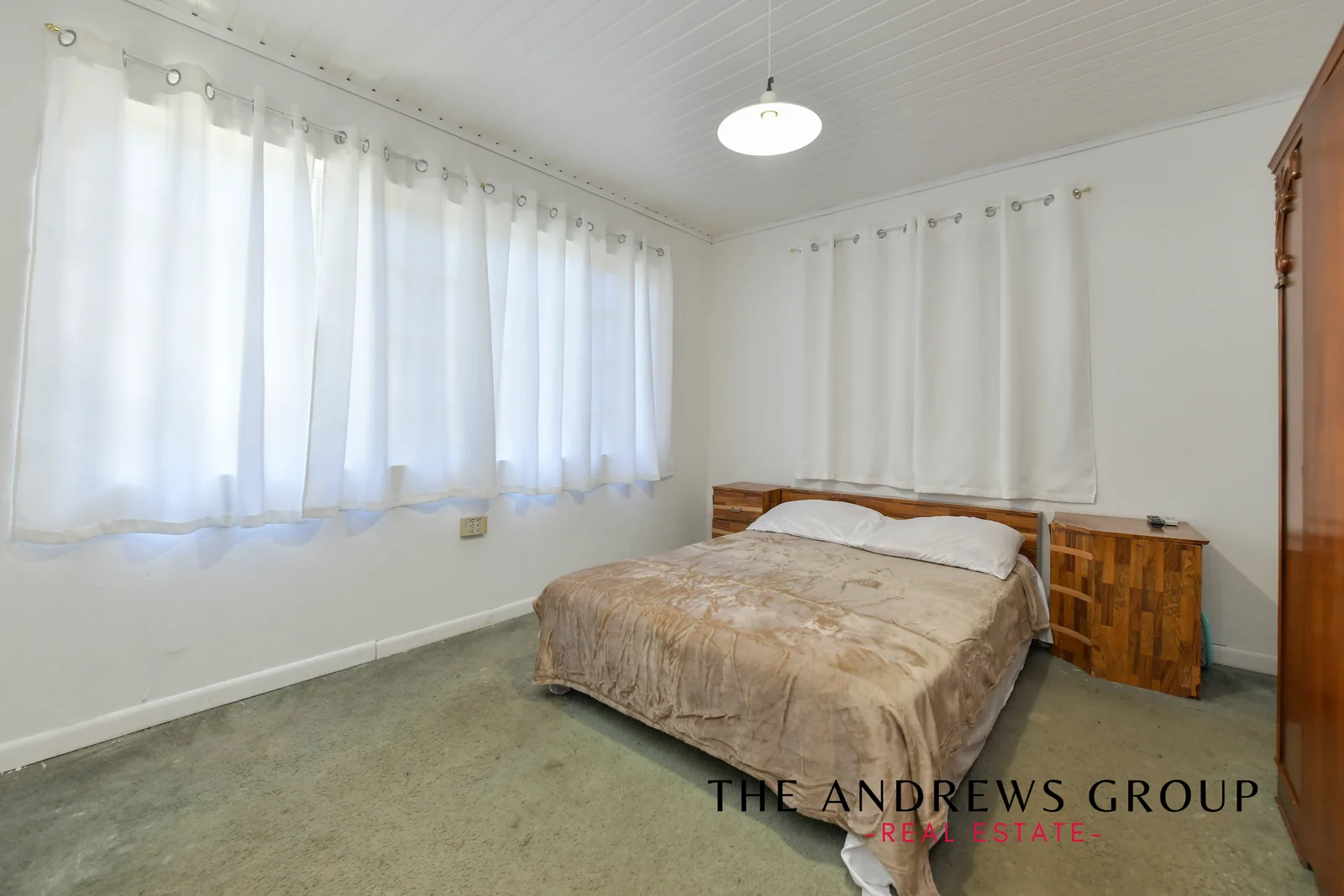 A bedroom with white walls, pale green carpet, a wooden bed, and two windows with white curtains.