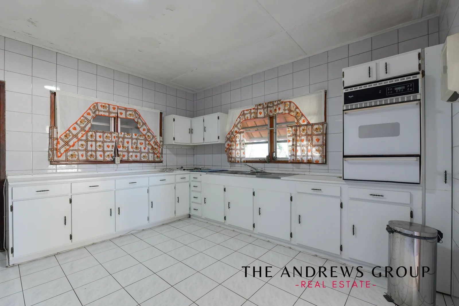 The main house kitchen: white tiled walls and floor, a long run of white cabinetry, a built-in wall oven, gas hob and stainless sink under two windows.