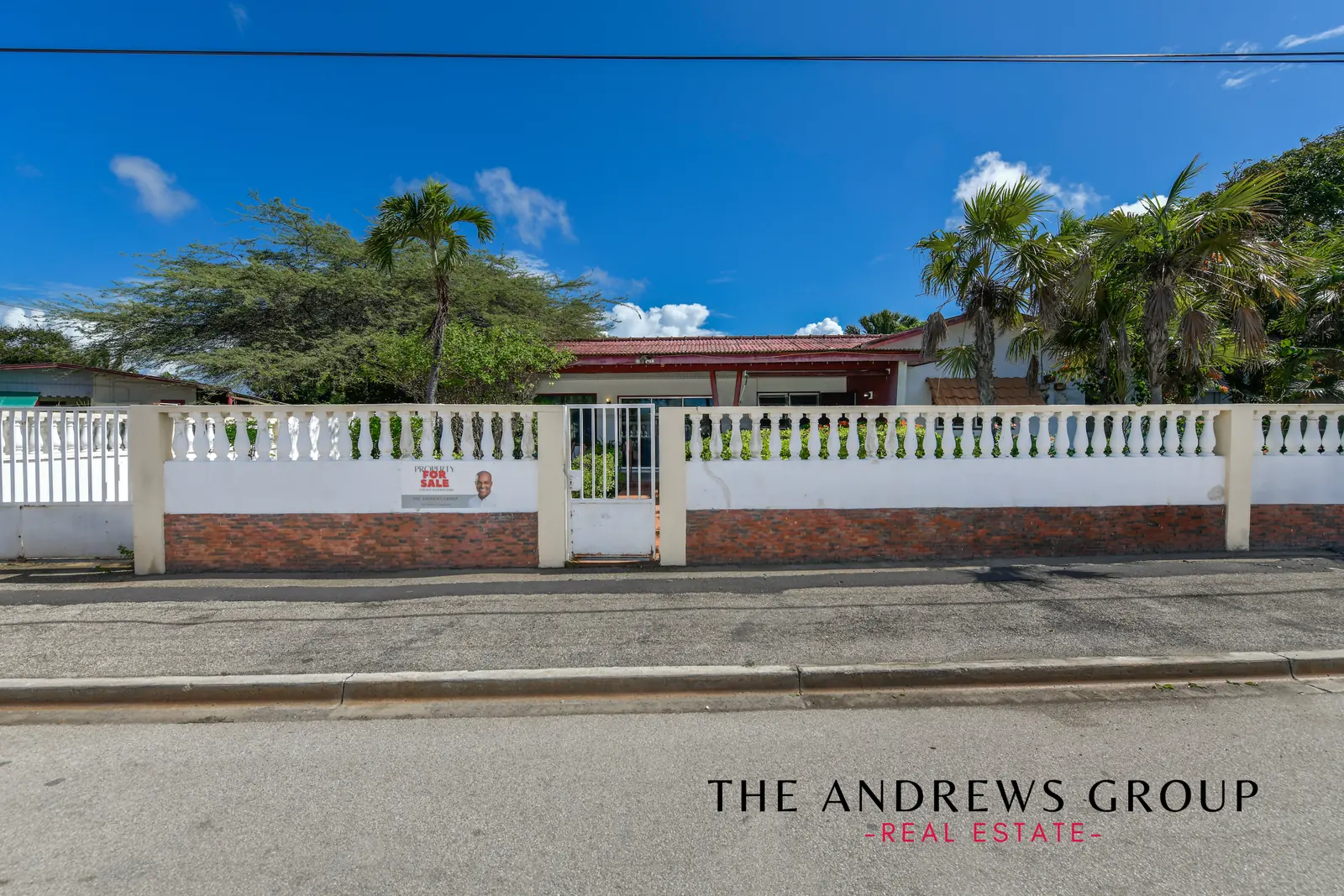The property seen from the road: a white balustrade wall and brick base, a pedestrian gate, palms, and the red-roofed veranda behind.
