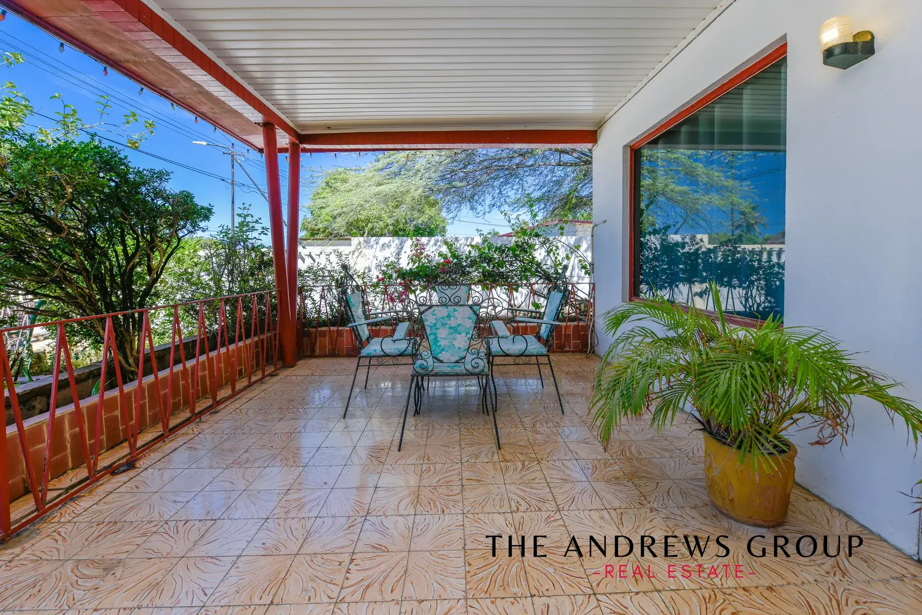 The veranda: patterned tile floor, red posts and railing, a wrought-iron table with turquoise cushions, potted palms, and the street through the greenery.